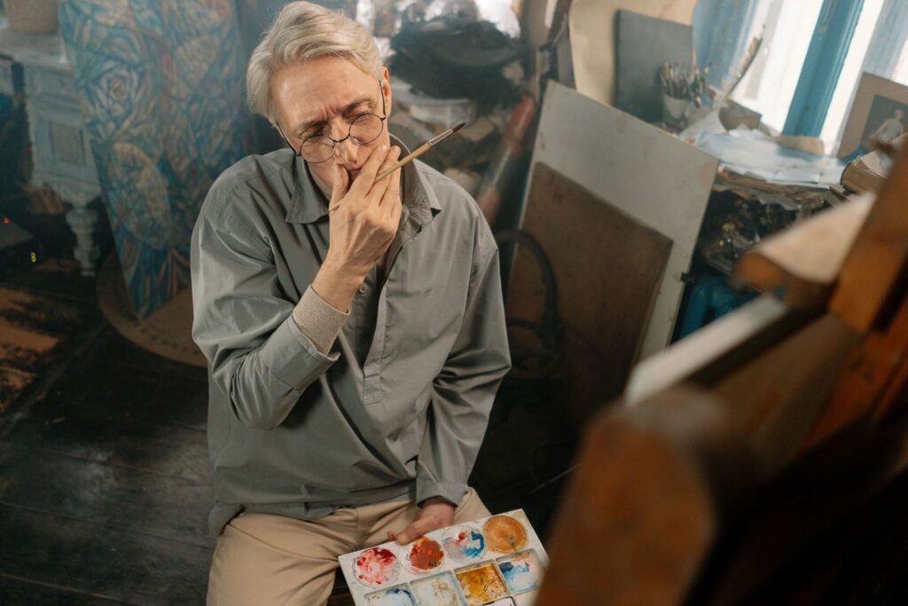 Older artist studying a canvas with brush and paint palette in hand